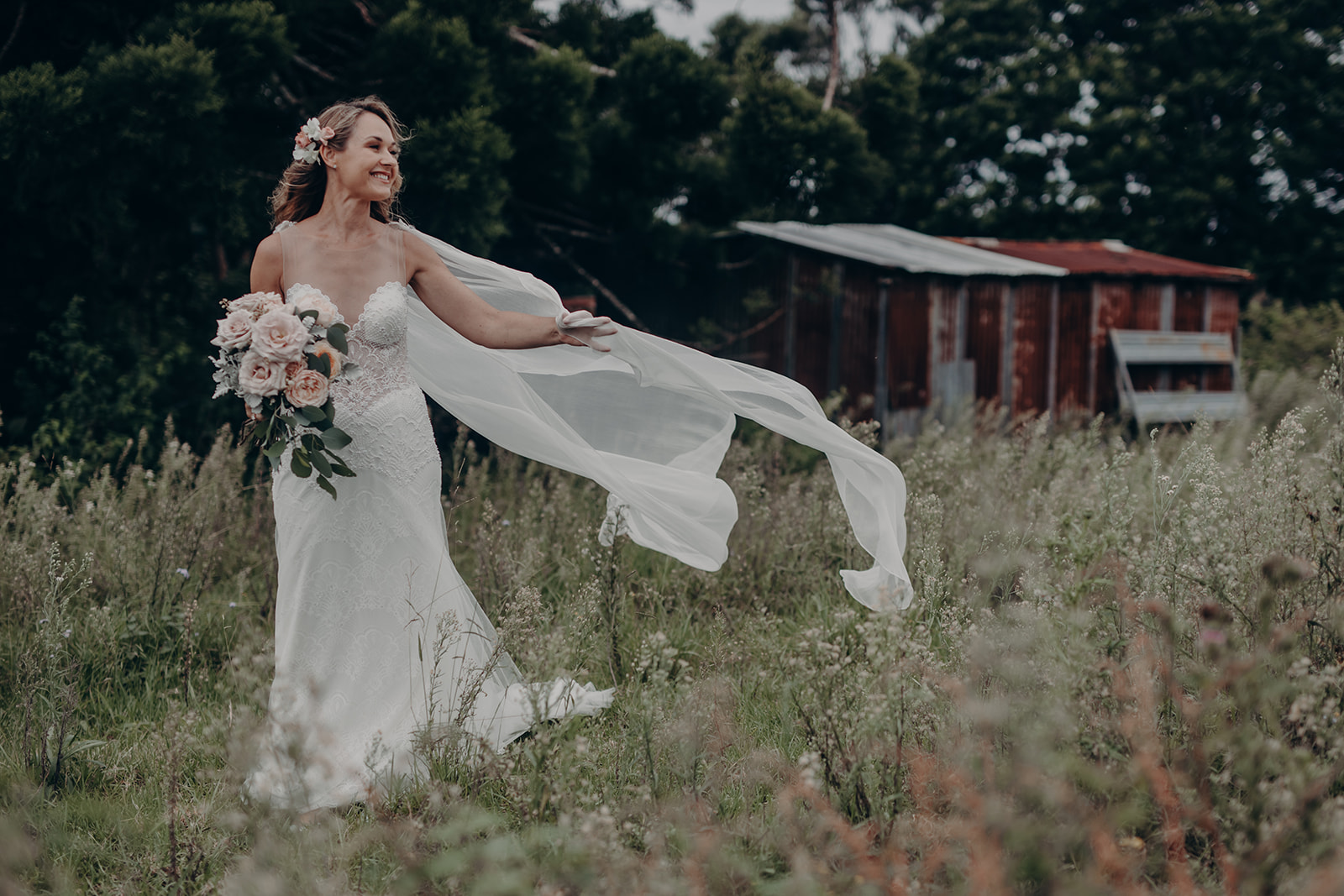A bride with a bouquet and a veil captured by the wind walking through the paddock at Aurora House