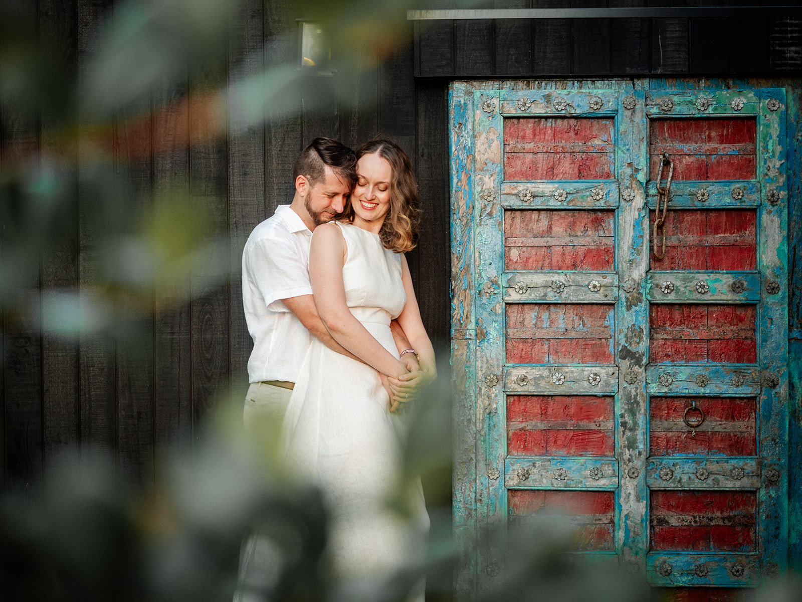 A newly engaged couple having an engagement shoot in front of a black wall with a red and green balinese door.